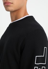 Sudadera negra con cuello redondo, con un ribete blanco y detalle de logo en la manga izquierda. Hecha de algodón con una textura suave.