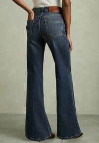 Jean évasé bleu foncé à taille haute, avec une patch en cuir portant le nom 'REISS' à l'arrière, des coutures doubles et deux poches arrière.