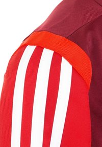 Bordeaux atletiekshirt met rode en witte gestreepte accenten op de schouder. Gemaakt van gladde stof met een geribbelde textuur op de mouwen.