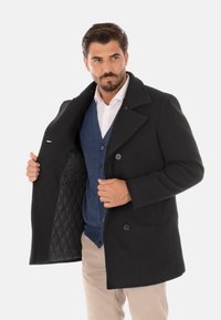 Cappotto lungo di lana nera con design doppiopetto, grandi bottoni e interno imbottito. Indossato sopra un cardigan blu.