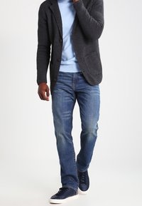 Mörktexturerad cardigan, ljusblå tröja, rakskurna blå jeans och marinblå sneakers med vita sulor. Funktioner inkluderar fickor på cardiganen.