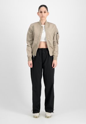 Beige bomber bunda se zipem vpředu, elastickými manžetami a cargo kapsou, spárovaná s černými kalhotami s elastickým pasem a bílými teniskami.