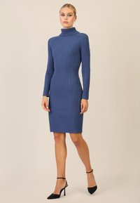 Abito lavorato a coste blu con collo alto e maniche lunghe, silhouette aderente, lungo fino al ginocchio, abbinato a scarpe con tacco alto nere.