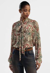 Blusa di seta floreale color crema con motivi rossi e blu, maniche lunghe e un dettaglio a fiocco sul collo. Abbinata a jeans di denim scuri.