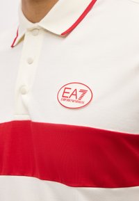 Άσπρο πόλο μπλουζάκι με κόκκινη οριζόντια ρίγα στο στήθος και γιακά με κόκκινη επένδυση, με το λογότυπο EA7 Emporio Armani στην αριστερή πλευρά.
