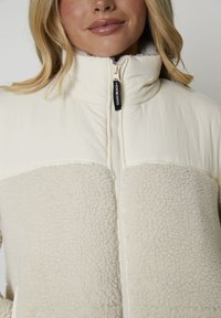 Mujer vistiendo una chaqueta beige claro con cremallera, con textura de forro polar en la parte inferior y tela suave en el pecho superior y el cuello.