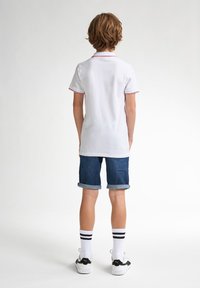 Valkoinen polo-paita punaisilla yksityiskohdilla, farkkushortsit, valkoiset urheil socksit mustilla raitoja, ja mustavalkoiset lenkkarit. Takaviisto.