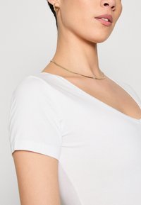 Un haut blanc à manches courtes et col en V met en valeur un tissu doux et une coupe ajustée. Un collier courbé en ton or et des boucles d'oreilles en créole apportent des accents.