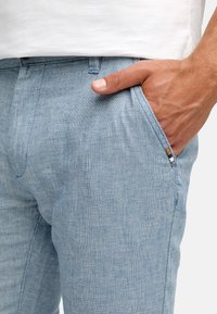 Hellblaue Hose mit Fischgrätmuster und einer Vordertasche, die eine kleine braun-weiße Akzentierung nahe der Tasche aufweist. Glatte Stoffstruktur.