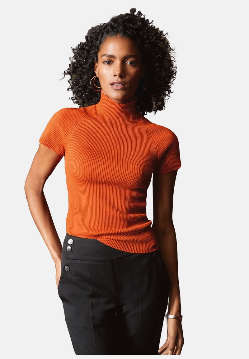 Korte mouwen, geribbelde coltrui in felle oranje. Gecombineerd met zwarte broeken met decoratieve zilveren knopen in de taille.
