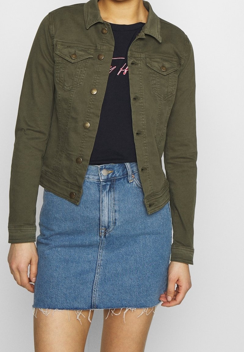 Dunkel olivgrüne Denimjacke mit Knopfverschluss, Brusttaschen und leicht verkürztem Design, getragen über einem schwarzen T-Shirt und einem blauen Jeansrock.