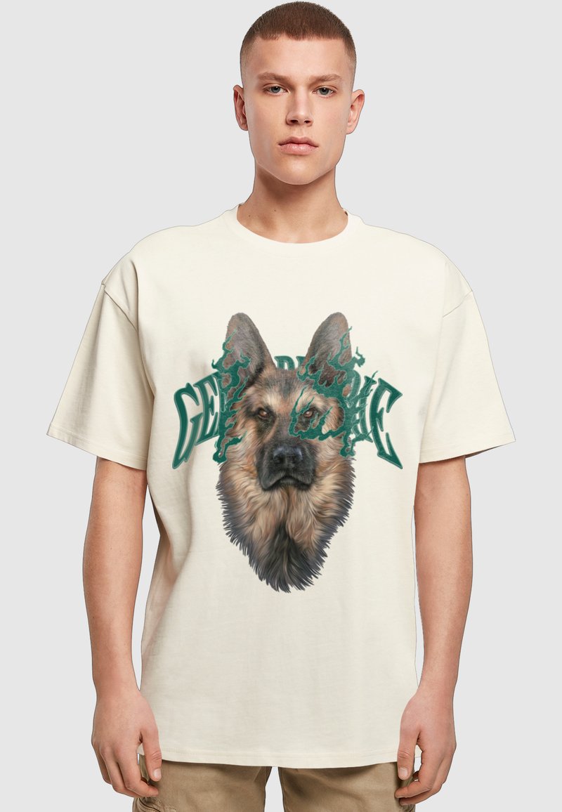 Νέος άνδρας που φοράει λευκό T-shirt με λεπτομερές πρόσωπο Γερμανικού Ποιμενικού και πράσινο κείμενο στο μπροστινό μέρος, στέκεται μπροστά σε απλό φόντο.