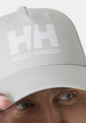 White Helly Hansen cap with large "HH" logo worn by person adjusting the brim, showing part of their eyes and forehead.