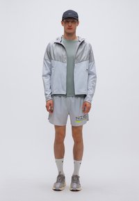 Αθλητικό μπουφάν ανοιχτού γκρι χρώματος με γυαλιστερή υφή, σετ με αντίστοιχο σορτς και πράσινη κοντομάνικη μπλούζα. Αξεσουάρ περιλαμβάνουν μαύρο καπέλο και κάλτσες.