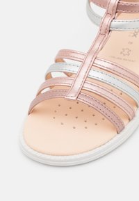 Sandaia de dedo abierta con tiras metálicas en rosa y plata, una plantilla suave en beige con perforaciones y una suela blanca.