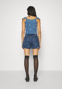Kék bordázott crop top virágmintával, magas derekú farmer rövidnadrággal és combközépig érő fekete harisnyával párosítva. Fekete lábbeli látható.