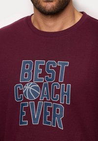 Maroon cotton t-shirt featuring blue text "BEST COACH EVER" with a basketball graphic, crew neck design, and short sleeves.