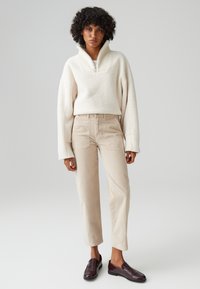 Pullover in tessuto crema con colletto alto a coste, pantaloni in velluto a coste beige e scarpe slip-on di un borgogna scuro. Design semplice con una vestibilità comoda.