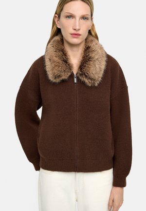 Kobieta w brązowym swetrze zapinanym na zamek z dużym, puszystym futrzanym kołnierzem i białych spodniach, stojąca na jednolitym tle.