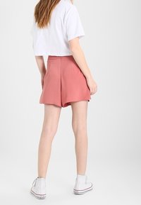 Oříznuté bílé tričko ve spojení s světle růžovými šortkami. Šortky mají hladkou texturu a volný střih, končící uprostřed stehna. Bílé tenisky.