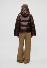 Pruun kapuutsiga puffer-jakk, millel on läikiv, ruuduline tekstuur ja laiad varrukad, sobitatud beežide laiade pükstega.