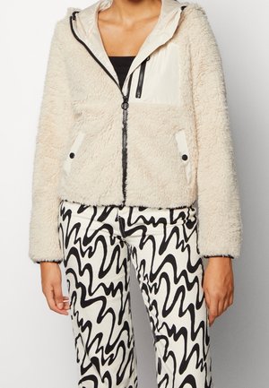 Giacca in finta pelliccia beige chiaro con cappuccio crema liscio, chiusura a zip e tasche laterali, abbinata a pantaloni a righe bianco e nero.