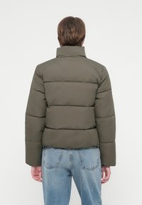 Person iført en kort olivengrøn pufferjakke og lyseblå jeans, vendt væk mod en simpel hvid baggrund.