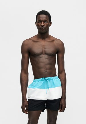 Atletisk mørkhudet mand stående iført badebukser i blå, hvid og sort farveblokering mod en ensartet lys baggrund.