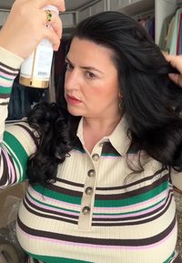 Une femme aux longs cheveux noirs applique un produit capillaire d'un flacon pulvérisateur. Elle porte un t-shirt à rayures multicolores à côtes avec des boutons.