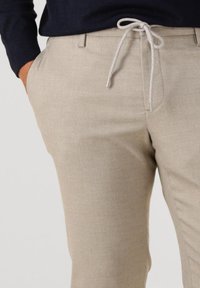 Beige herringbone pants with a drawstring waist. Fabric is smooth with a subtle texture; features side pockets and a tailored fit.