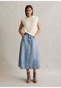 Lichtcrème gebreide mouwloze top, gecombineerd met een blauwe denim maxi rok, met een uitlopende snit en voorzakken. Bruine enkellaarsjes maken de look compleet.