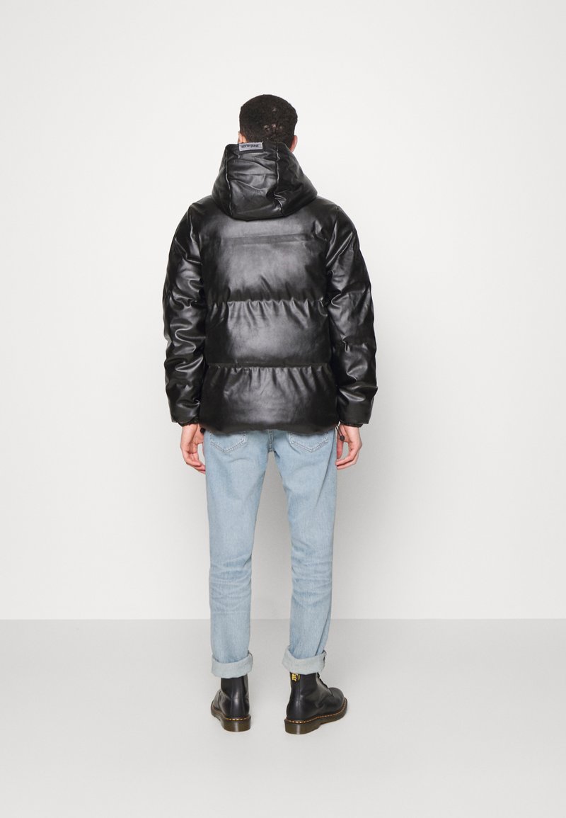 Doudoune noire avec une finition brillante et lisse, dotée d'une capuche et de sections matelassées horizontales. Portée sur un jean bleu clair et des bottes.