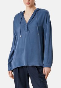 Haut à capuche bleu à manches longues avec encolure en V, doté d'une patte de boutonnage et d'un cordon de serrage. Tissu doux et lisse ; coupe décontractée.