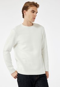 Junger Mann mit kurzen, hellbraunen Haaren, der ein weißes, längs geripptes Langarmshirt und dunkle Jeans trägt. Er blickt zur Seite, mit den Händen in den Hüften.