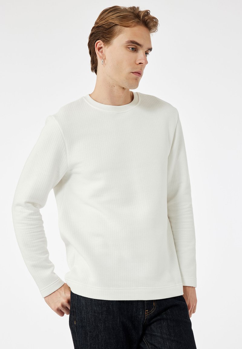 Ung mand med kort lysebrunt hår iført en hvid ribbet langærmet skjorte og mørke jeans, som ser til siden med hænderne på hofterne.