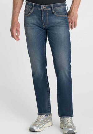 Uomo che indossa jeans di denim blu e scarpe da ginnastica argentate, in piedi davanti a uno sfondo chiaro e uniforme.