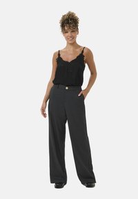 Schwarzes Spitzen-Camisole mit dünnen Trägern, kombiniert mit hoch taillierten, weit geschnittenen grauen Hosen mit einem goldenen Knopfverschluss.