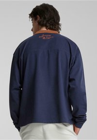 Marineblå langermet skjorte med oransje krage. Har brodert tekst på baksiden. Laget av glatt stoff; løs passform med enkelt design.