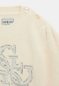 Maglione crema con colletto a costine, impreziosito da filo argento e paillettes in un logo stilizzato. Presenta due bottoni decorativi chiari.