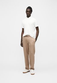 Homme debout portant un polo blanc à manches courtes, un pantalon beige avec cordon de serrage et des baskets blanches sur un fond clair uni.