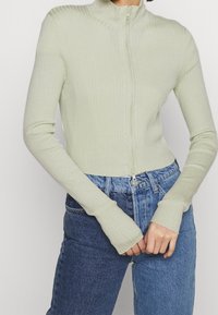 Person klädd i en ljusgrön ribbad zip-up tröja med tumhål och tvåfärgad blå högmidjade jeans, stående framför en enfärgad bakgrund.