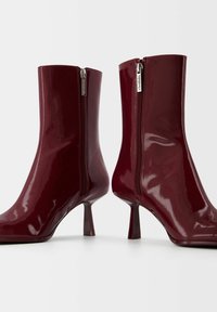 Bottes cheville rouges foncées brillantes avec fermetures éclair latérales et talons carrés sur une surface blanche.