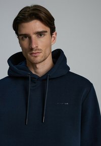 Granatowy hoodie wykonany z miękkiego materiału, z kapturem na sznurki, subtelnym brandingiem i gładką fakturą bez widocznych wzorów.