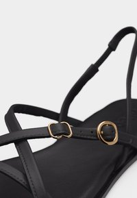 Μαύρο σανδάλι με λεπτές λουρίδες και δύο μικρές χρυσές αγκράφες σε λευκό φόντο.