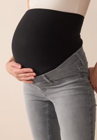 Crna rastezljiva elastika preko istaknutog trudničkog trbuha, u kombinaciji s sivim uskim trapericama koje imaju prednji džep i uski kroj.