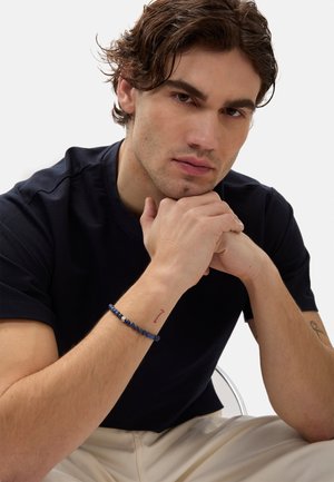 T-shirt navy a collo giro, vestibilità comoda. Primo piano di una mano con un braccialetto di perle blu e un piccolo tatuaggio rosso sul polso. Sfondo chiaro.