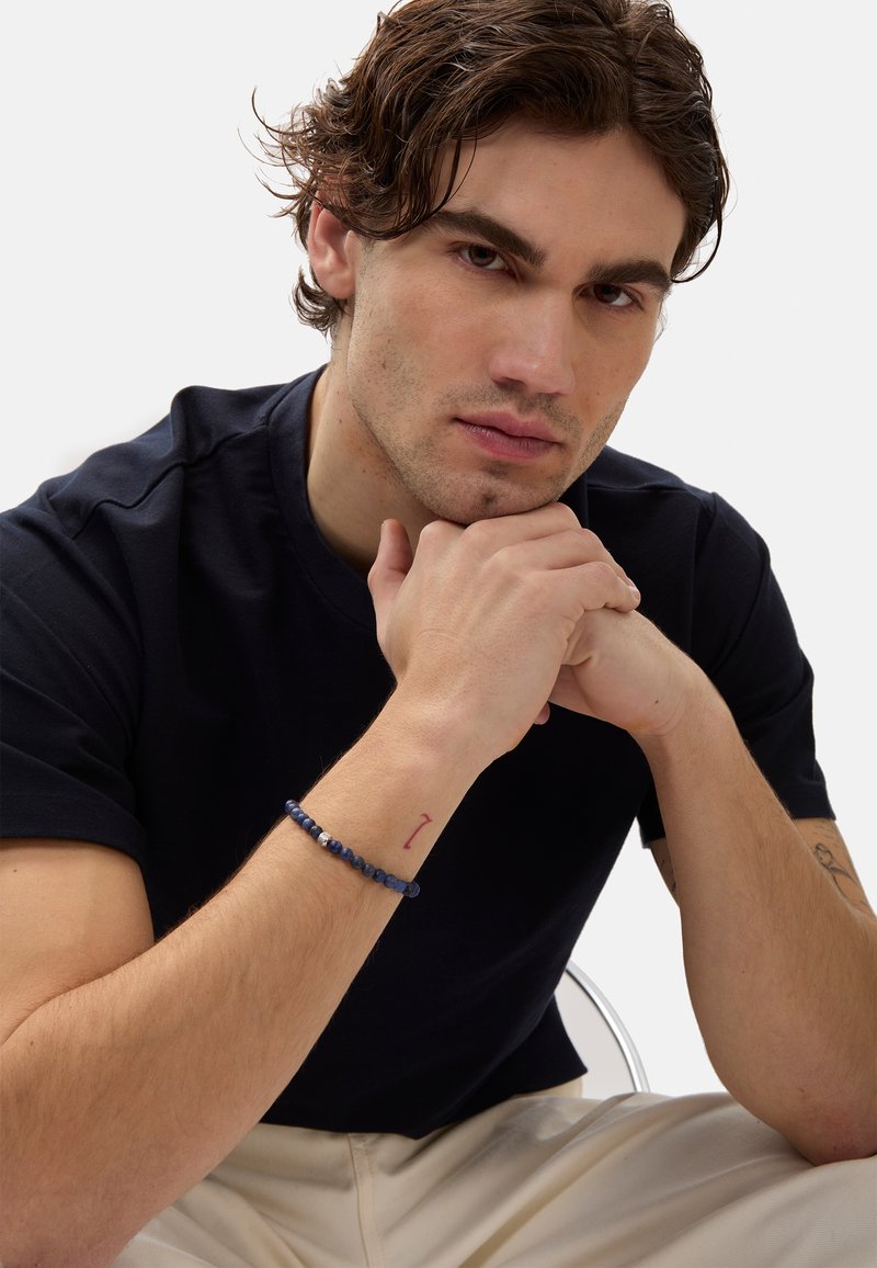 Marinblå crew-neck t-shirt, avslappnad passform. Närbild av en hand med ett blått pärlarmband och en liten röd tatuering på handleden. Ljus bakgrund.