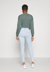 Sudadera verde recortada con mangas anchas, combinada con vaqueros ajustados azul claro con bajo deshilachado. Zapatillas blancas completan el conjunto.