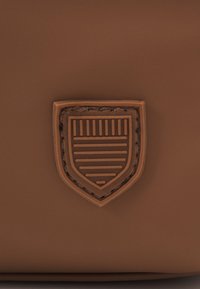 Surface en cuir brun avec une pièce écusson en forme de bouclier cousue, présentant un motif linéaire au centre.