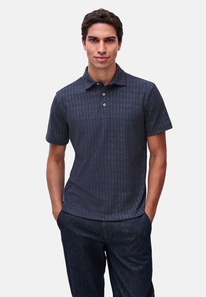 Hombre con una camiseta polo azul oscuro de manga corta con un patrón de rayas verticales texturizadas y pantalones oscuros, de pie con las manos en los bolsillos.